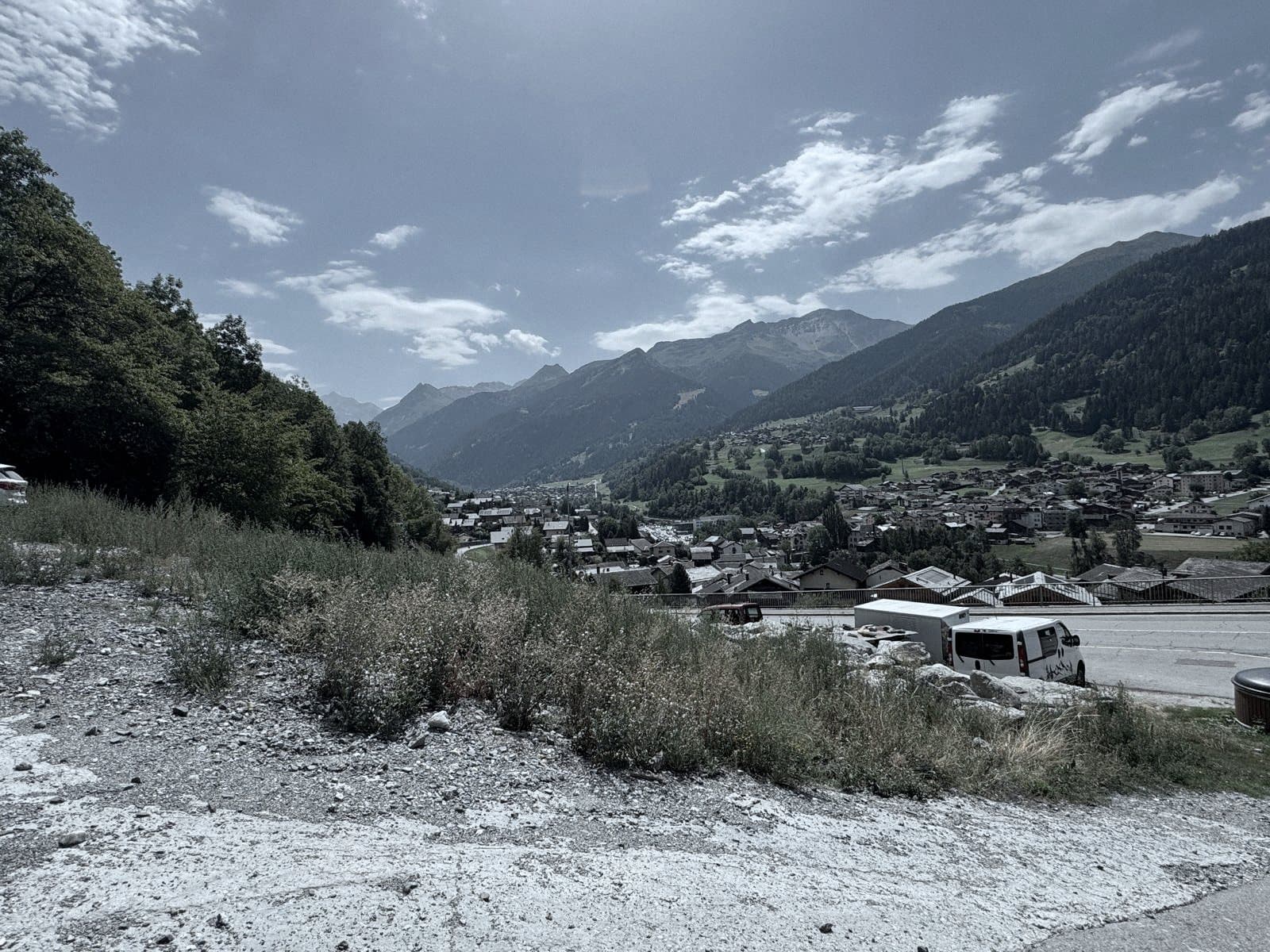 Un terrain libre au-dessus de la route, herbes sèches et gravier, la vallée et le village en arrière-plan.
