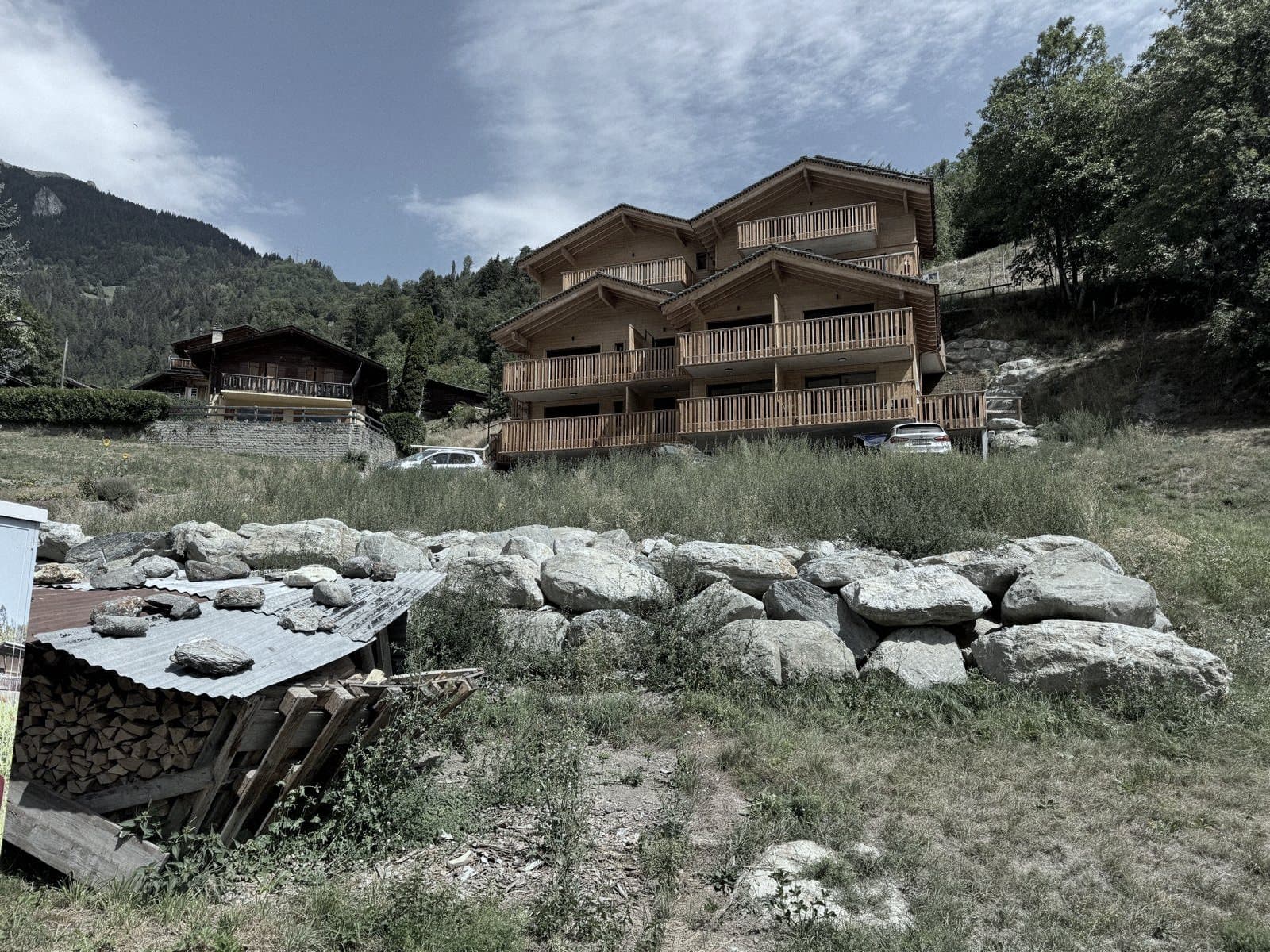 Une résidence récente en bois à balcons filants, adossée à la pente à côté de chalets plus anciens.